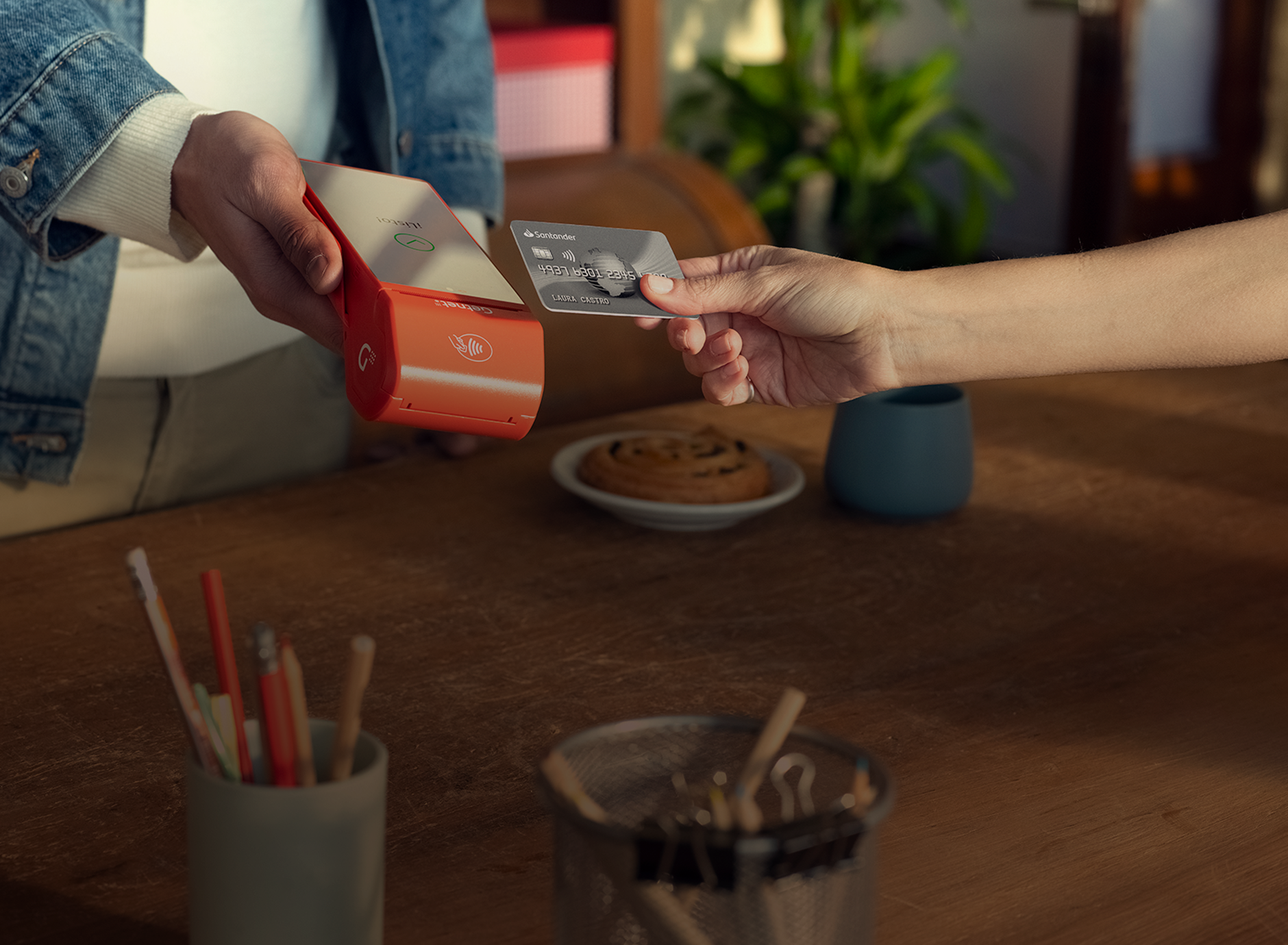 Mano sosteniendo una terminal roja de Getnet mientras otra acerca una tarjeta para pago sin contacto, sobre una mesa con lápices y una taza.