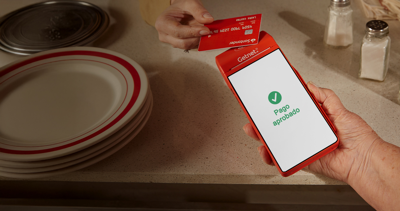 Mano sosteniendo la terminal roja de Getnet con pantalla mostrando “Pago aprobado”, mientras otra mano acerca una tarjeta sobre un mostrador.