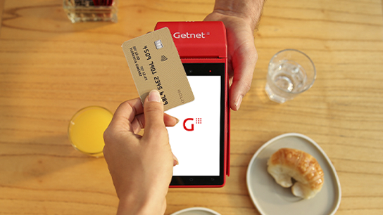 Persona realizando un pago con tarjeta en una terminal roja de Getnet sobre una mesa con desayuno.