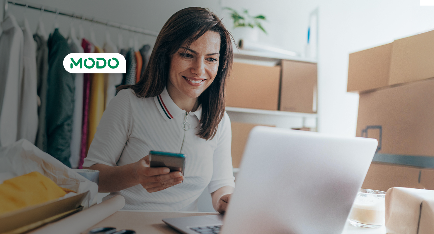 Persona usando laptop y teléfono móvil en un espacio con cajas y perchero con ropa, sobre escritorio con objetos.