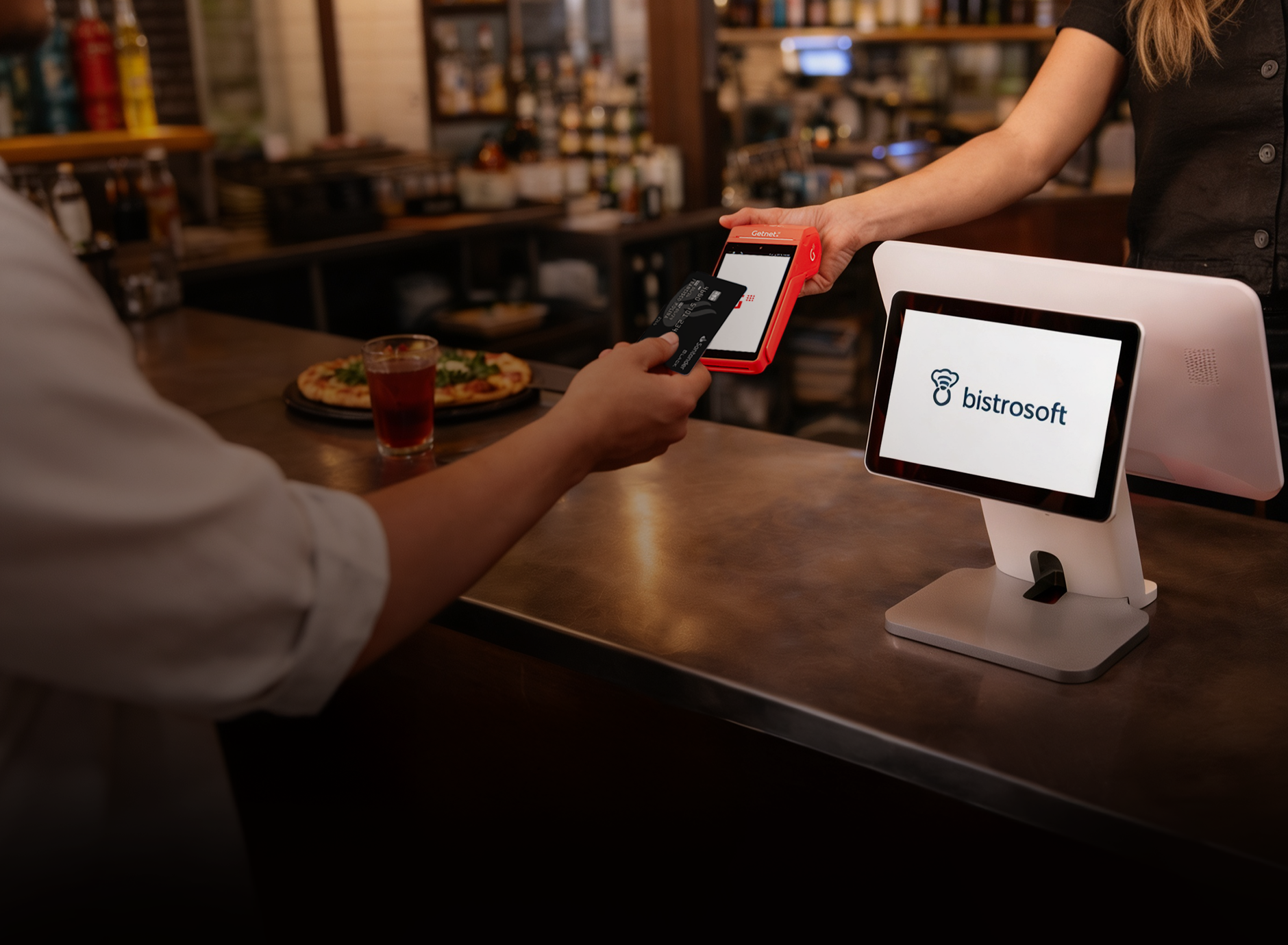 Pago con tarjeta apoyada sobre una terminal roja de Getnet, junto a una pantalla de sistema de gestión Bistrosoft en un mostrador.