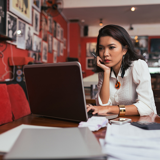 Persona usa una laptop sobre mesa con papeles en un café, con decoración de cuadros en el fondo.
