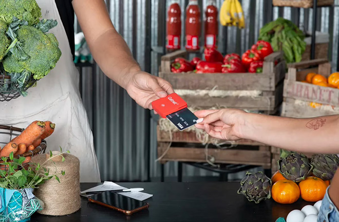 Persona insertando una tarjeta en un lector rojo de Getnet sostenido por otra persona, sobre un mostrador con verduras y frutas en un mercado.